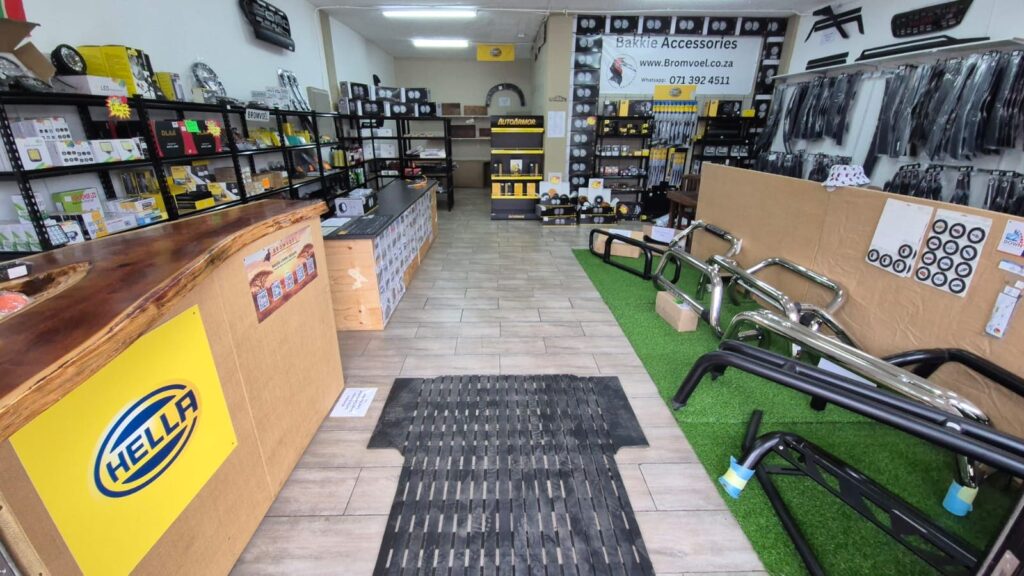 Inside a car parts store: a wooden counter on the left, tall shelves with accessories, a 'Bakkie Accessories' banner overhead, and metal vehicle racks on a green display area to the right.