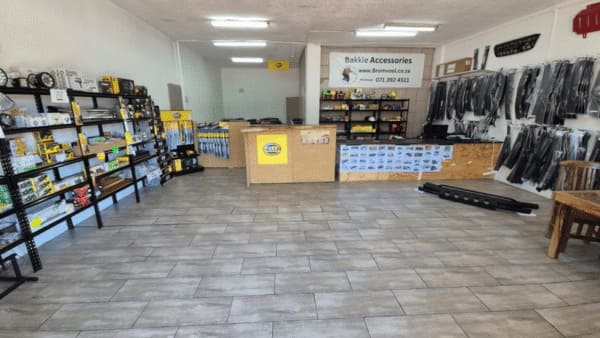 Interior of a shop with auto parts on shelves along the left and right walls, a central checkout counter, and a 'Bakkle Accessories' sign at the rear wall.